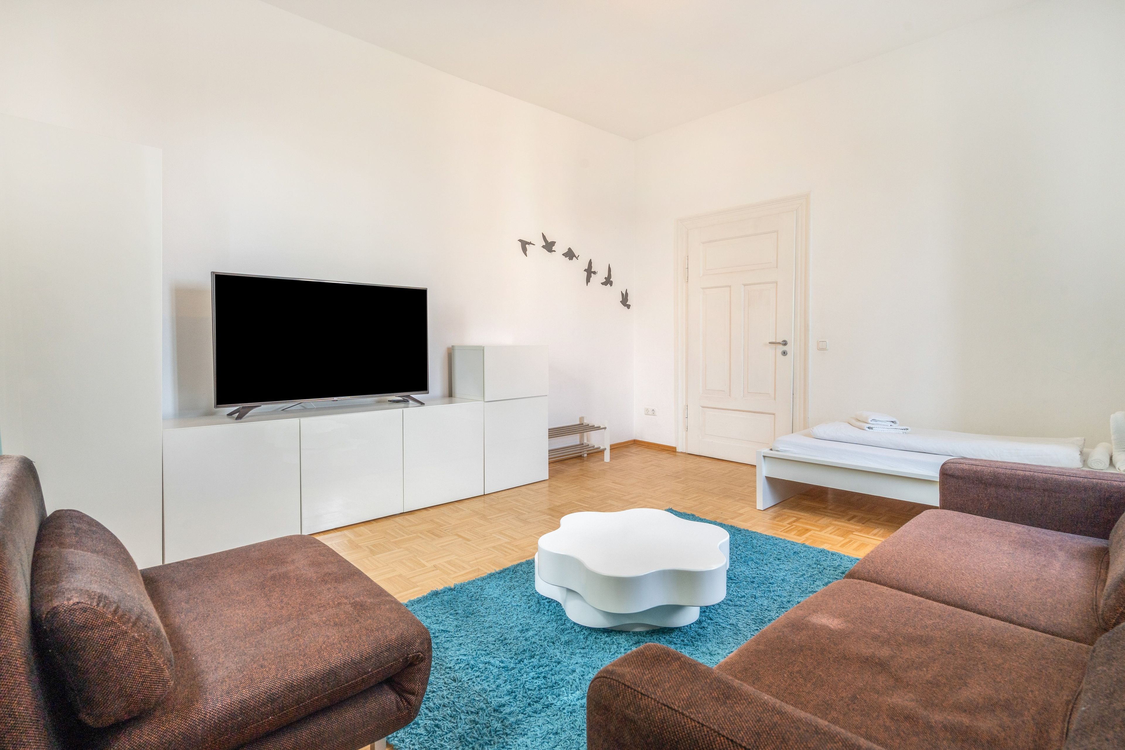 Una sala de estar moderna con un televisor de pantalla plana, muebles blancos, un tapete azul y una puerta con manija.