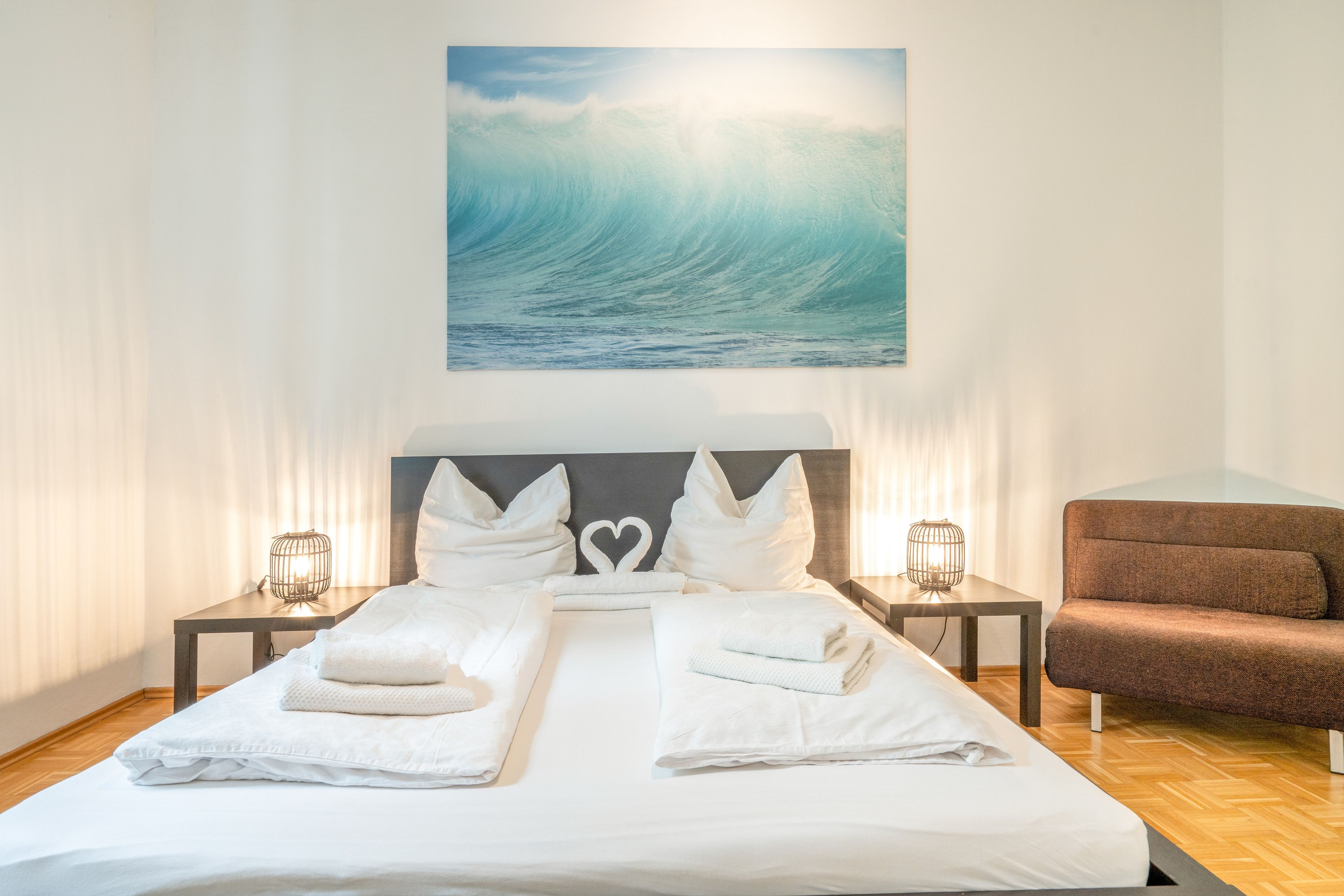 Une chambre d’hôtel avec un grand lit, deux tables de chevet avec des lampes, un fauteuil brun et un tableau représentant une vague, accroché au mur.
