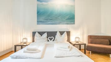 Une chambre d’hôtel avec un grand lit, deux tables de chevet avec des lampes, un fauteuil brun et un tableau représentant une vague, accroché au mur.