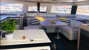 Interior de un yate con comedor, asientos, una pequeña cocina y una planta en maceta.