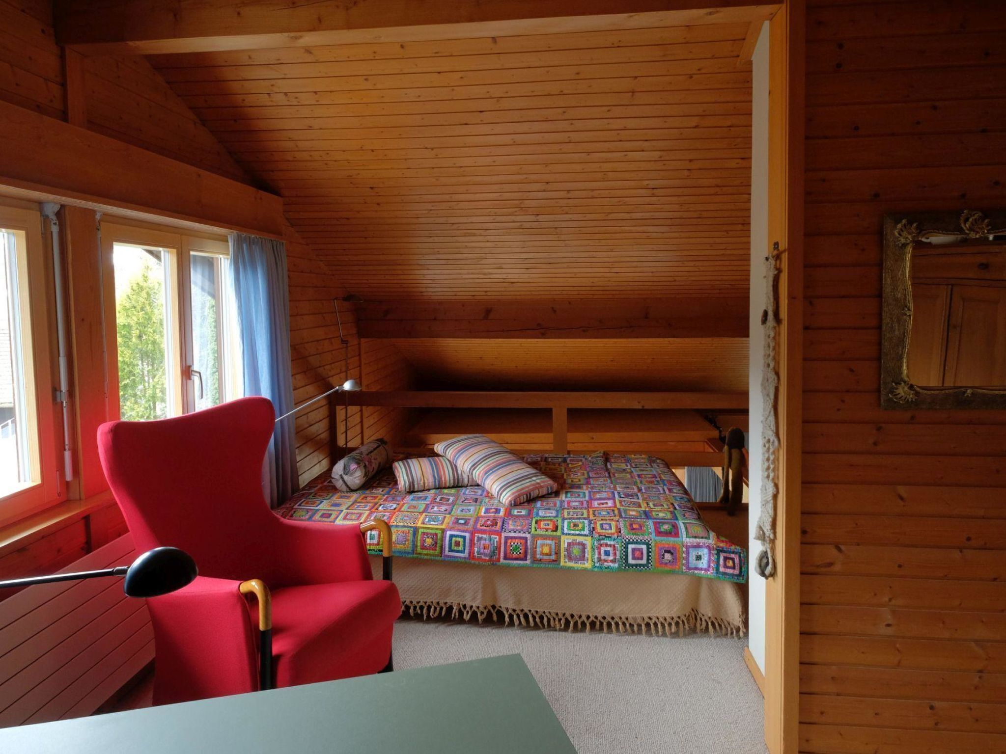 Une chambre confortable dans un chalet, avec un plafond en bois, un lit simple recouvert d’une courtepointe aux couleurs vives, une chaise rouge et une table.