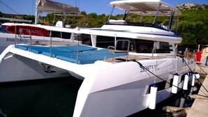 Una barca bianca ormeggiata in una marina, con un ponte blu, un bozzello di salvataggio e una struttura che funge da cabina.