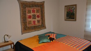 Een slaapkamer met een bed, een groot wandtapijt, een lijst met een afbeelding en een bureau met een lamp.