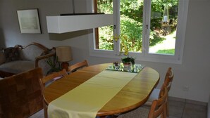Een eethoek met een houten tafel, stoelen, een gele tafelkleed, een bank, een lamp en een raam met uitzicht op groen.