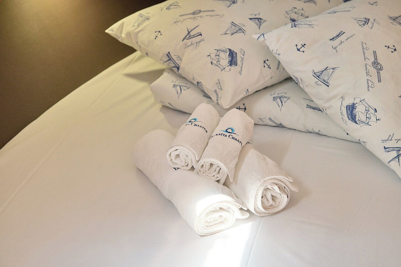 Ein ordentlich gemachtes Bett mit weißen Leinen und zusammengerollten Handtüchern, ein Kissen mit nautischem Muster und ein Kissen mit Aufschrift.