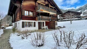 Un chalet de madera de dos pisos con un balcón, un patio cubierto de nieve y montañas al fondo.