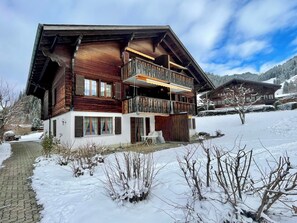Eine zweistöckige Holzhütte mit Balkon, einem verschneiten Hof und einem Berg im Hintergrund.