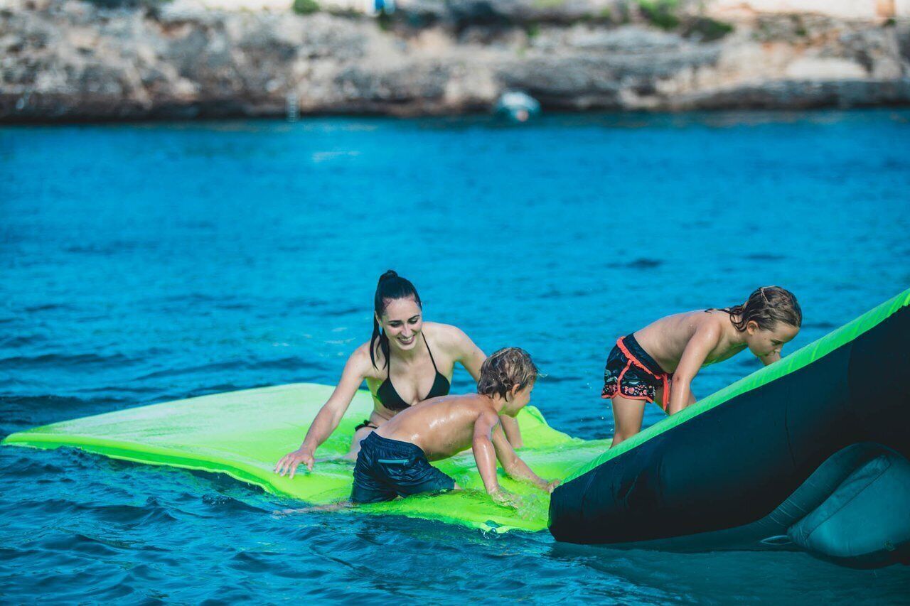 Three people on a green inflatable raft in a body of water with a rocky shoreline in the background.
