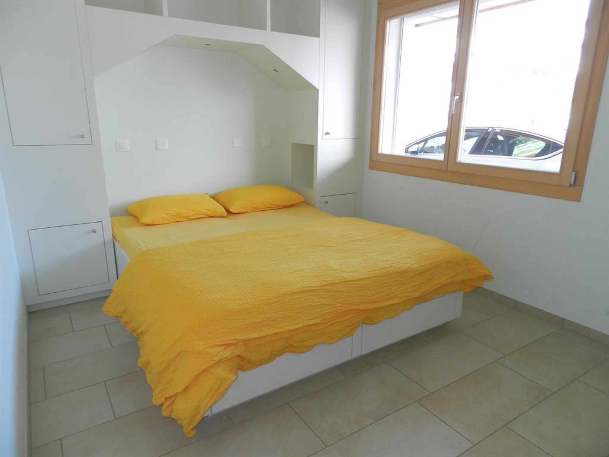 Un lit simple avec une couvre-lit jaune, dans une pièce aux murs blancs et dotée d’une fenêtre.