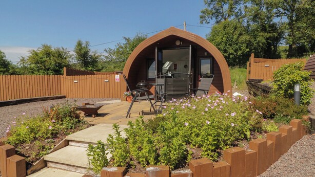 Een kleine, gedeeltelijk open constructie met een gebogen dak, een terras met buitenmeubilair en een tuin met planten en bloemen.