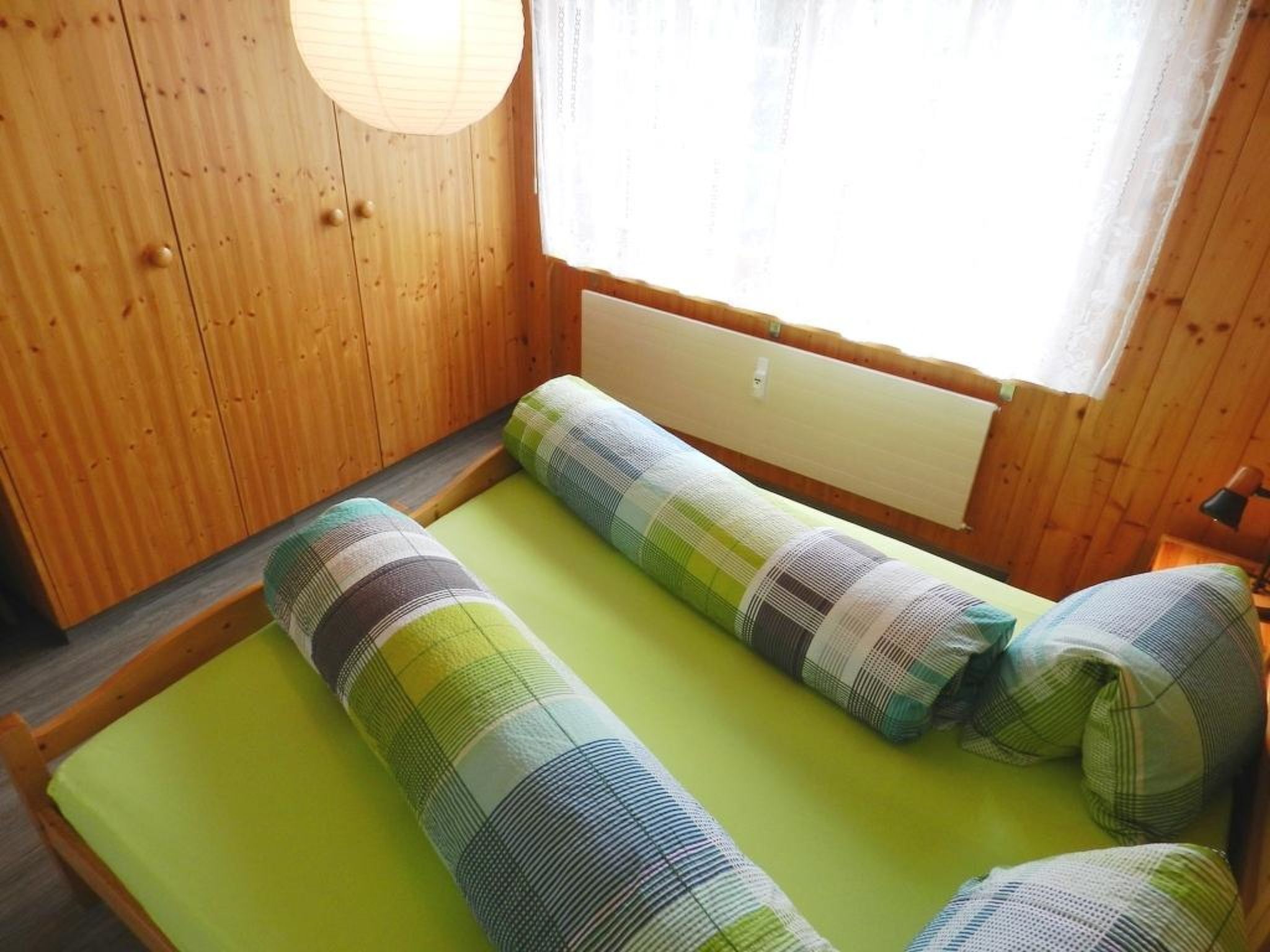 Huone, jossa on puinen sisustus, vihreällä patjalla varustettu sänky ja raidallinen vuodevaatteisto sekä ikkuna, jossa on verhot.