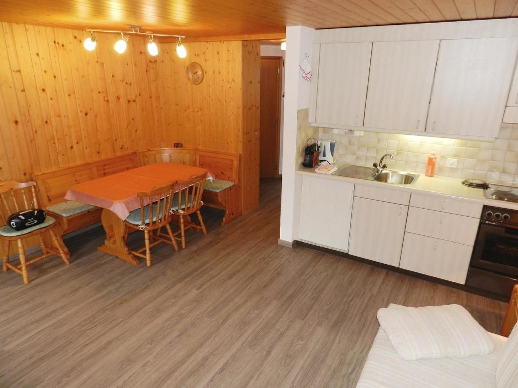A compact room with a wooden dining table, chairs, a kitchenette, and a bed with a white blanket.