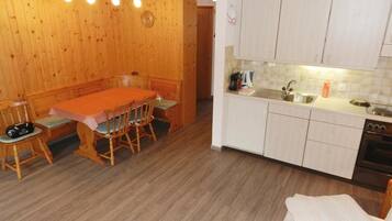 A compact room with a wooden dining table, chairs, a kitchenette, and a bed with a white blanket.