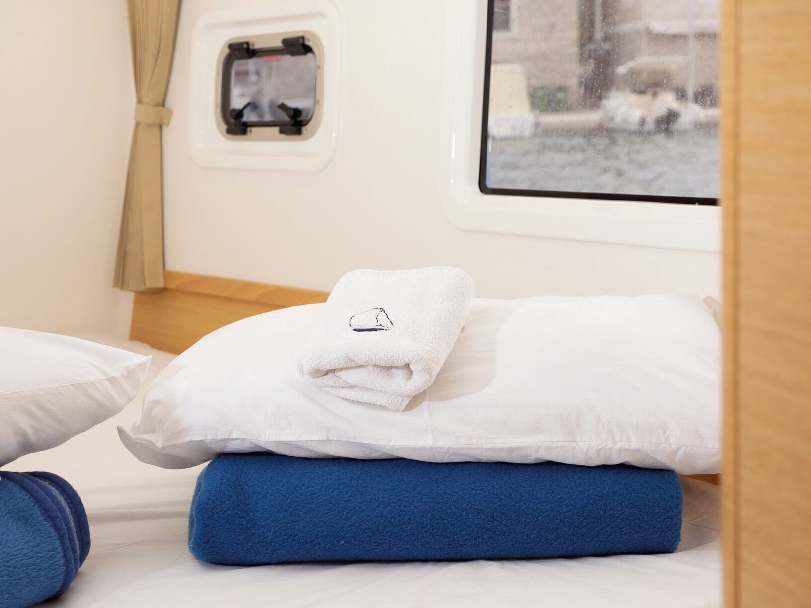 Una cama con ropa de cama blanca y azul, una ventana con vistas a un edificio, y un ventanal pequeño con un panel de control.