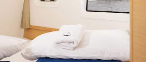 Una cama con ropa de cama blanca y azul, una ventana con vistas a un edificio, y un ventanal pequeño con un panel de control.