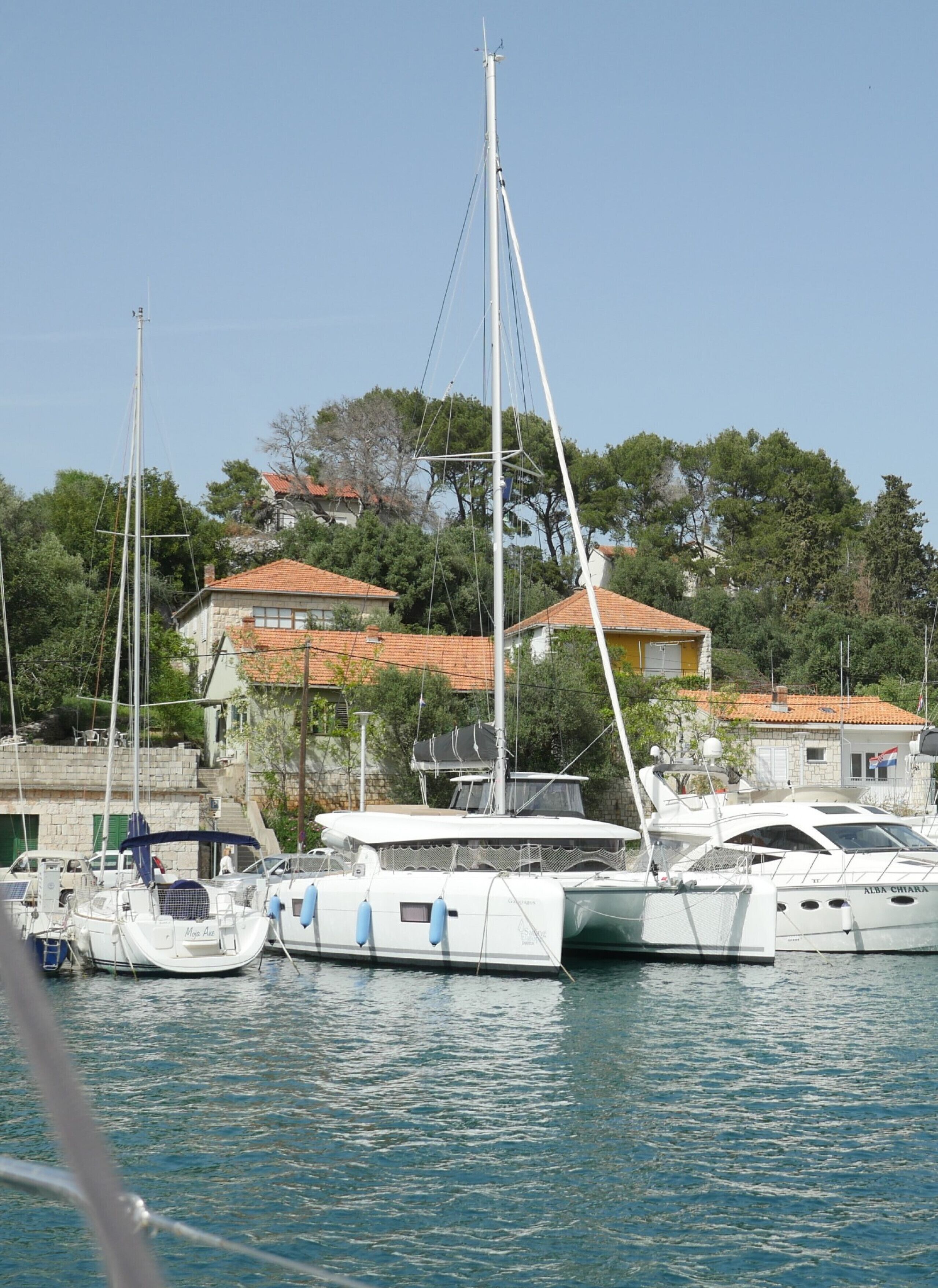 Uma marina com barcos atracados, um edifício de pedra com telhado de telhas vermelhas e árvores ao fundo.