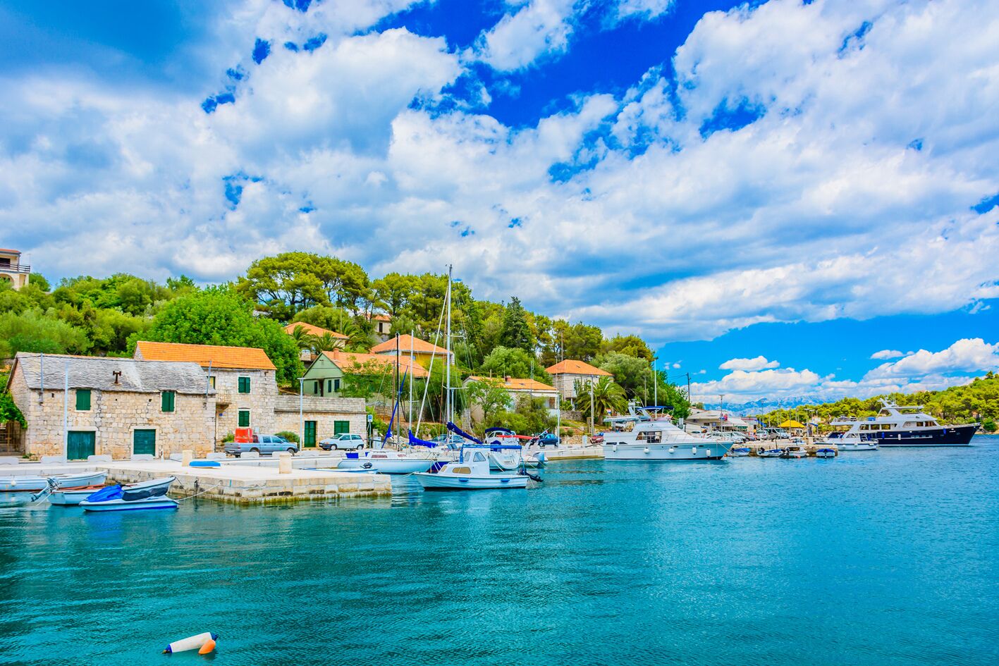 A coastal marina with boats docked, stone buildings with red roofs, and a clear blue sky.