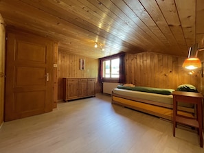 Una habitación con paredes y techo de madera, una cama con colchón verde, un armario de madera y una ventana con cortinas.