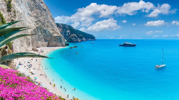 Uma praia costeira com águas turquesa cristalinas, um penhasco rochoso e um veleiro.