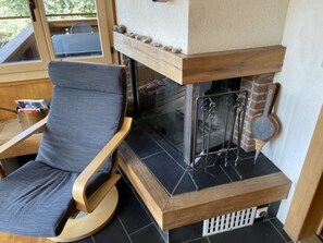 Une chaise à ossature en bois avec un coussin gris, une cheminée dotée d’une grille en métal et une fenêtre donnant sur un paysage verdoyant.