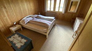 A small, single-bed room with a wooden ceiling, a radiator, a bedside table, a dresser, and a small table with a painting. - Sonnige Matte Appartement/Fewo, Dusche oder Bad, WC 8-Bettwohnung by Interhome (Gstaad)