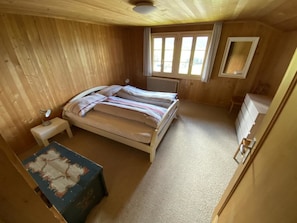 A small, single-bed room with a wooden ceiling, a radiator, a bedside table, a dresser, and a small table with a painting. - Sonnige Matte Appartement/Fewo, Dusche oder Bad, WC 8-Bettwohnung by Interhome (Gstaad)