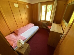 Een kleine eenpersoonskamer met een houten vloer, een radiator, een raam met gordijnen en een bureau met stoel.