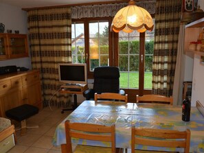 Ein Essbereich mit einem Tisch, der mit einer Tischdecke bedeckt ist, Holzstühle, einem Fernseher auf einem Schrank und einem Fenster mit Vorhängen.