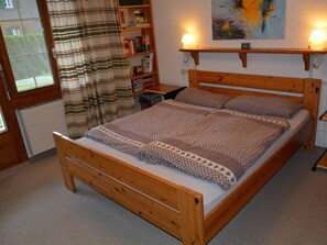 A wooden bed with a patterned bedspread and pillows, a bedside table with a lamp, a bookshelf with books, and a window with curtains.