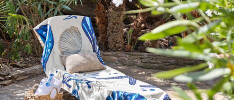 A sun lounger with a patterned cover, a cushion, and a woven basket on a stone patio surrounded by greenery.