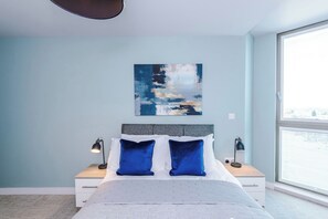 A bedroom with a bed, two bedside tables, a nightstand, a window, and a wall-mounted painting. - 1BDR Open Plan Apartment With Full Kitchen (Stretford)