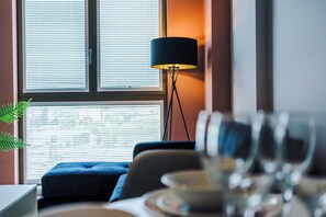 A dining area with a table set for two, a blue sofa, a floor lamp, and a window with blinds. - 1BDR Open Plan Apartment With Full Kitchen (Stretford)