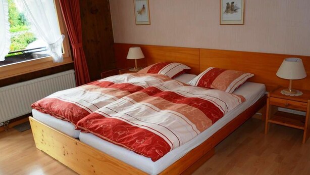 A wooden bed with a red and white bedspread, two bedside lamps, and framed pictures on the wall. - Mösli (Zimmerli) Appartement/Fewo, Dusche oder Bad, WC 2-Bettwohnung by Interhome (Zweisimmen)
