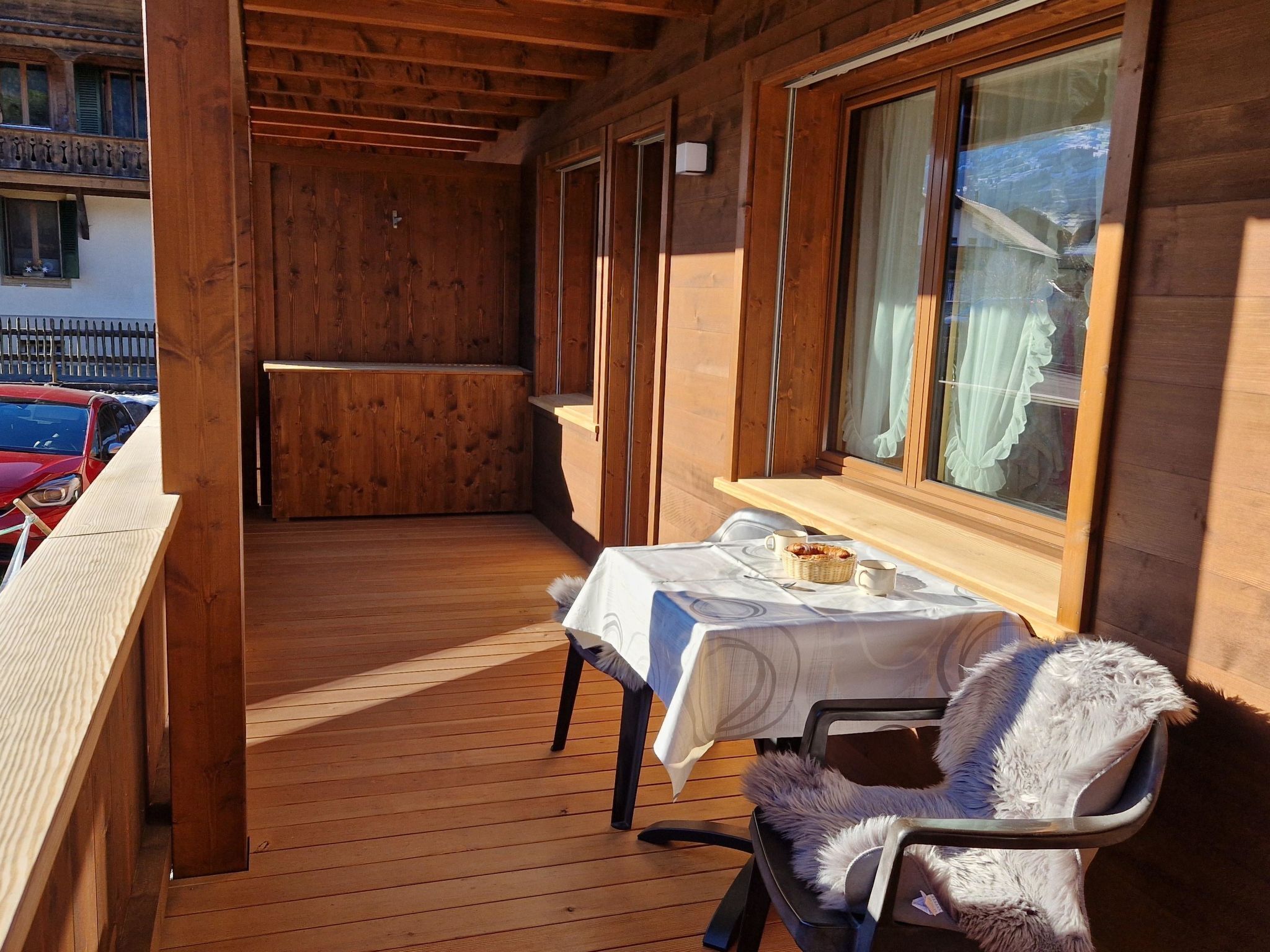 A wooden deck with a table and chair, a window with curtains, and a view of a building in the background.