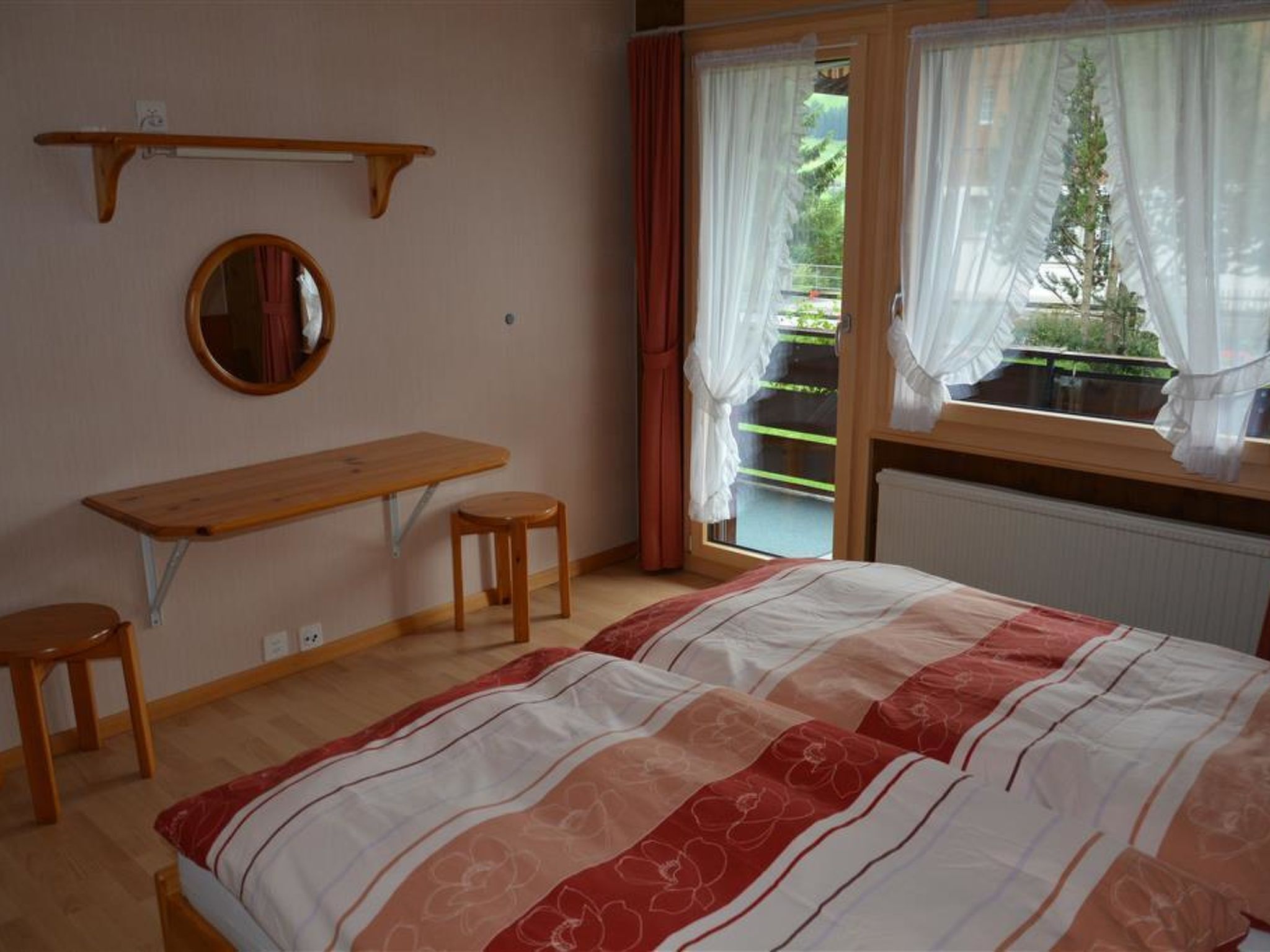 A bedroom with a bed, a desk, two stools, a mirror, and a window with curtains.