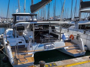 A white yacht docked at a marina with a covered deck area, a small table with chairs, and a lifebuoy. - Sun-Kissed Sailing Adventure in Islas Sarónicas (Alimos)