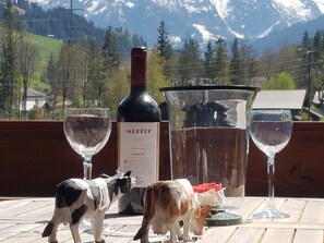 En flaska vitt vin från Mezzek, två vinglas och två små figurer föreställande kor, placerade på ett bord med utsikt över ett berg i bakgrunden.