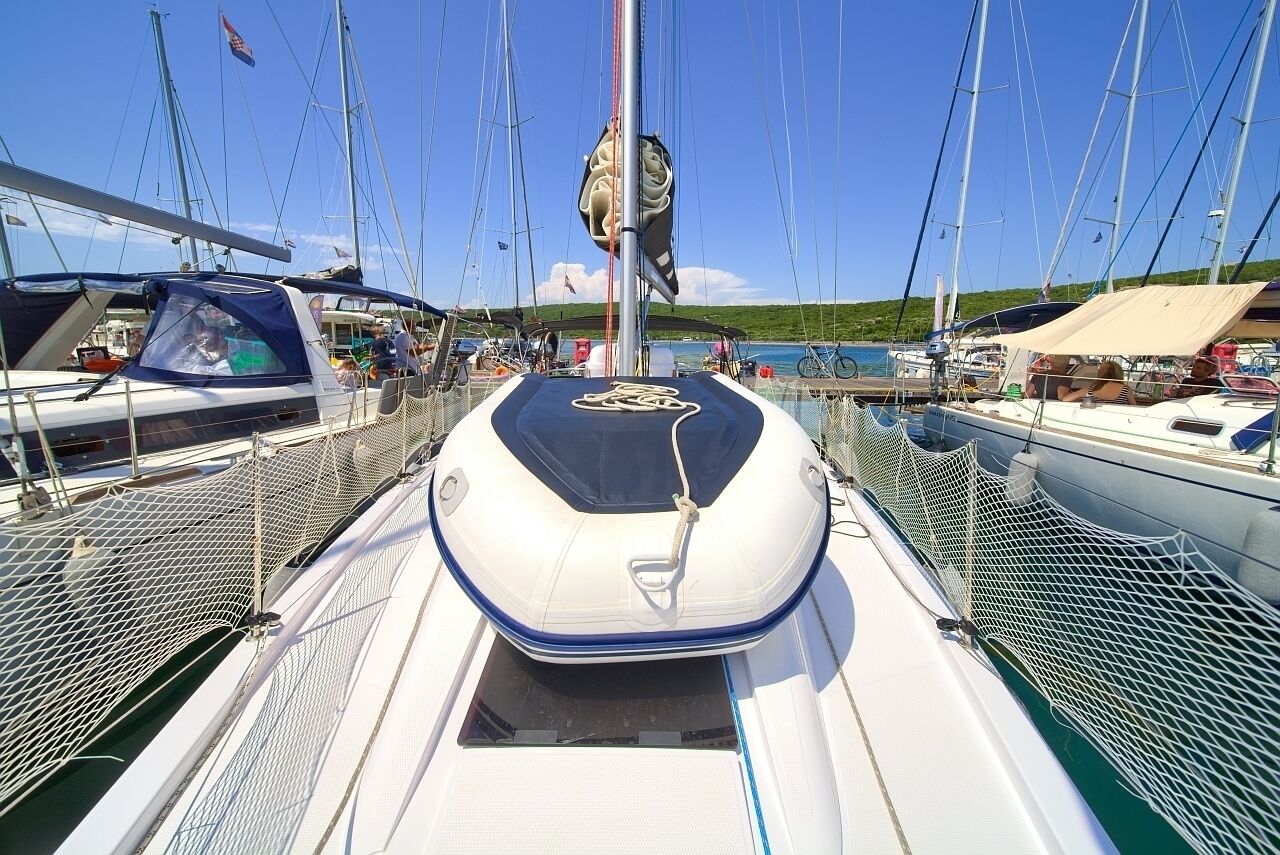 Un voilier est amarré dans une marina, avec un ciel dégagé, d’autres bateaux et un paysage verdoyant en arrière-plan.