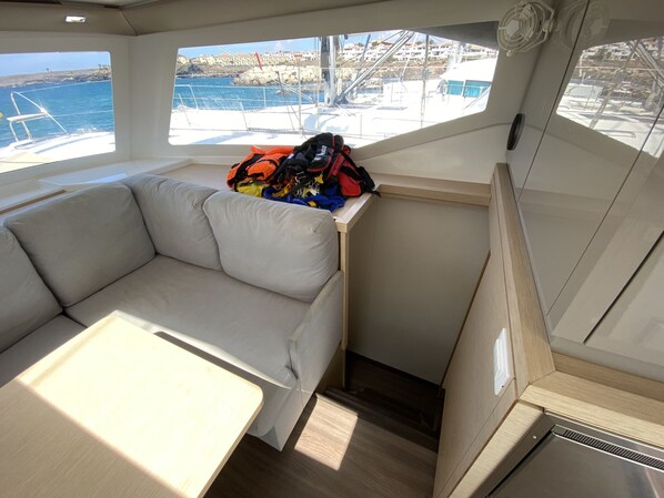 Interior de barco com área de estar acolchoada, uma mesa pequena e vista para a água e edifícios.