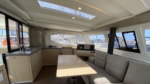 Eine kleine Kombüze mit Spüle, Herd und einem Tisch, eine gepolsterte Sitzecke und Blick auf den Hafen durch große Fenster.