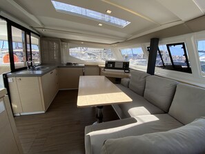 Eine kleine Kombüze mit Spüle, Herd und einem Tisch, eine gepolsterte Sitzecke und Blick auf den Hafen durch große Fenster.