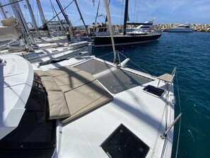 Un port de plaisance où sont amarrés de nombreux voiliers, un espace de repos couvert avec des coussins, et une digue rocheuse en arrière-plan.