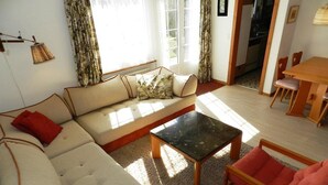 Una sala de estar con un sofá, una mesa de centro y una silla. Hay una ventana con cortinas, una puerta y una pequeña mesa con sillas.