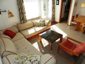A living room with a sofa, a coffee table, and a chair. There is a window with curtains, a door, and a small table with chairs. - Fink Appartement/Fewo, Dusche oder Bad, WC 2-Bettwohnung by Interhome (Zweisimmen)
