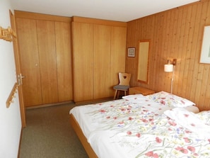 A bedroom with wooden walls, a bed with floral bedding, a chair, a bedside table, and a mirror. - Fink Appartement/Fewo, Dusche oder Bad, WC 2-Bettwohnung by Interhome (Zweisimmen)