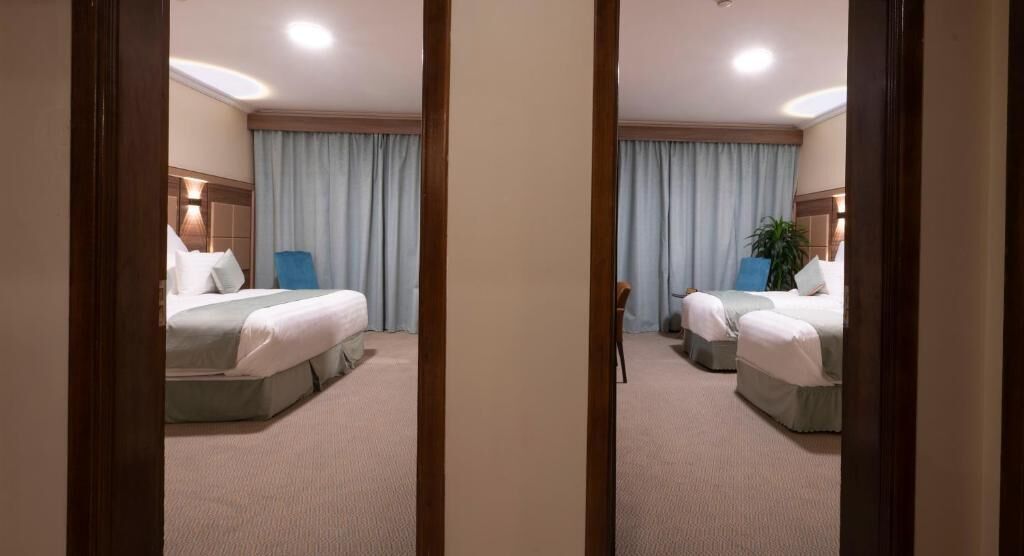Habitación de hotel con dos camas, un ventanal con cortinas, una mesita de noche y una lámpara de pared.