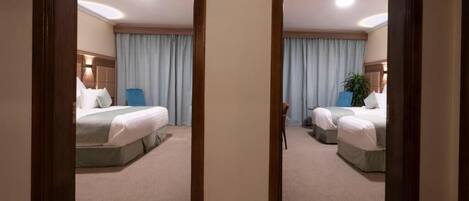 A hotel room with two beds, a large window with curtains, a bedside table, and a wall-mounted lamp.