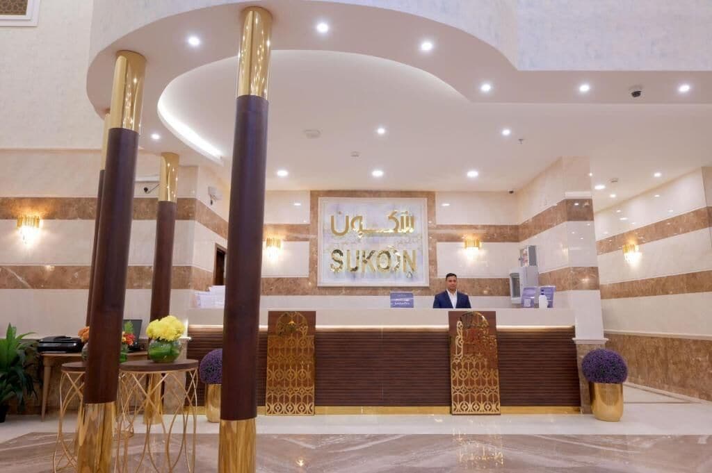 A hotel reception area with a modern design, featuring a reception desk, a man standing behind it, and decorative elements like plants and vases.
