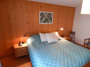 Ein Schlafzimmer mit einer Holzwand, einem Bett mit blauer Steppdecke, zwei Nachttischen mit Lampen und einem floralen Bild im Rahmen über dem Bett.
