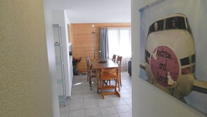 Een eethoek met een houten tafel en stoelen, een keuken met een koelkast en kasten, en een poster aan de muur.